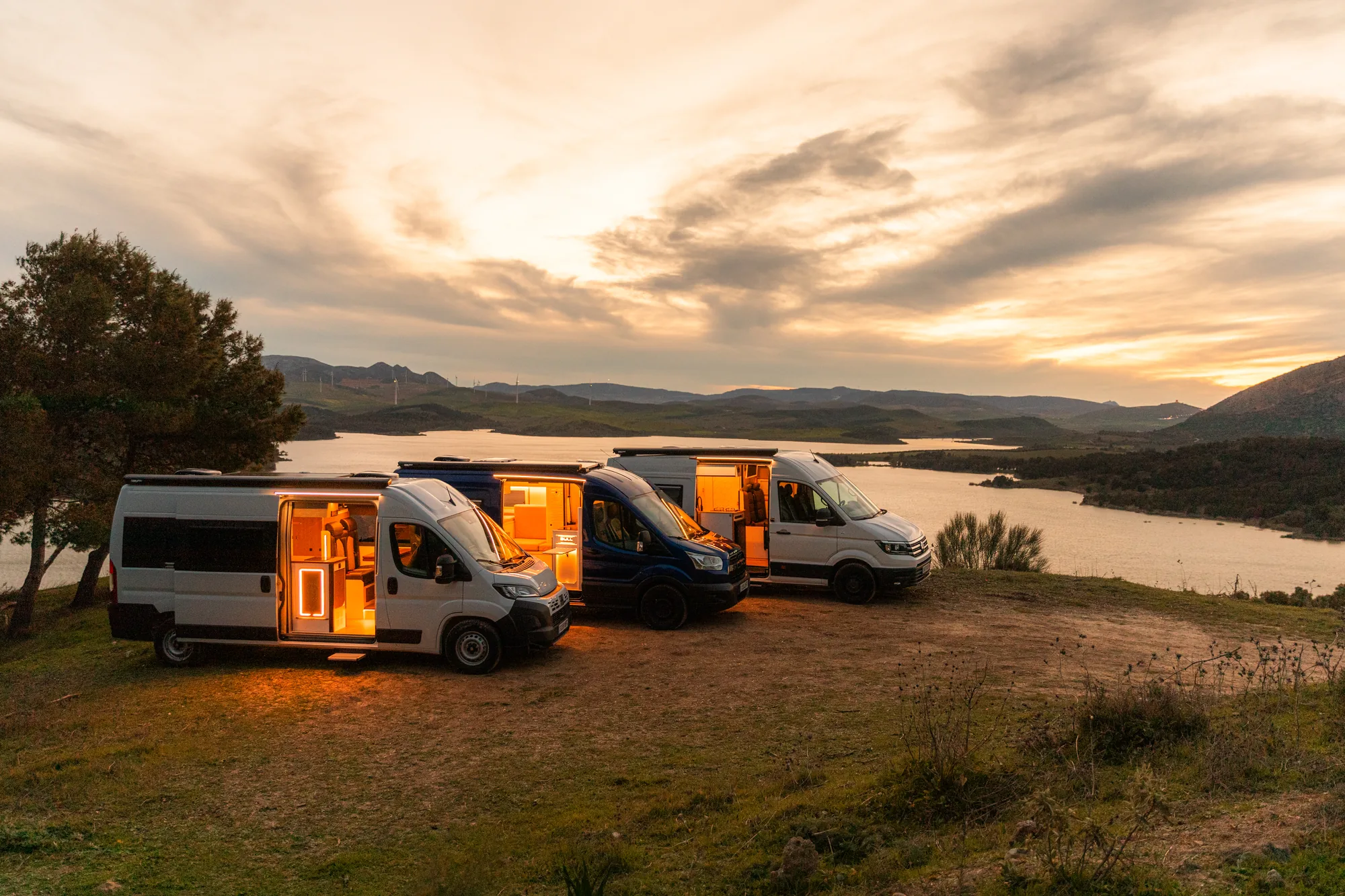 Tres furgonetas camperizadas Bullcamper con las puertas laterales abiertas junto a un embalse al atardecer